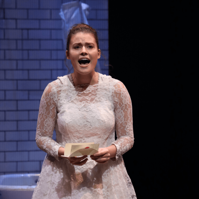 a young woman with brown hair wears a white lacy dress while singing on a stage