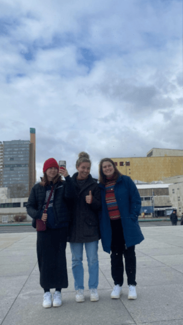 a group of three young women stand outside in winter clothes on a cloudy day