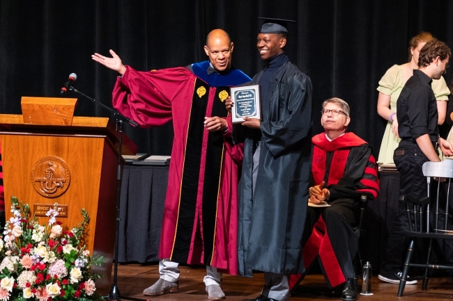 a young Black man accepts an award while wearing regalia on a stage