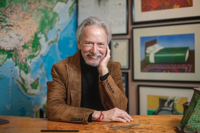 an older white man wearing a black top and jacket sitting in front of a wall of maps and prints