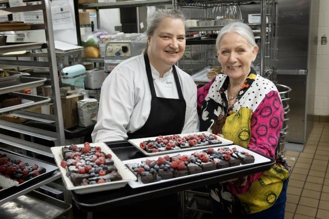 two middle aged women hold trays of food in a kitchen while smiling
