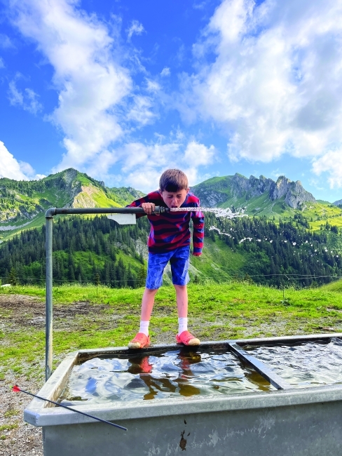 a young man takes a sip of water from a spigot in front of the Alps