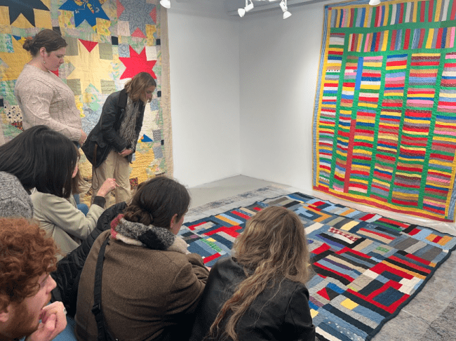 a group of students stand around examining a collection of quilts in an art gallery