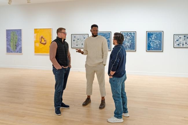 a group of three men stand together in an art gallery talking