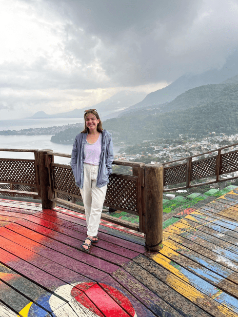 a young white woman standing on a wooden deck overlooking mountains and a river