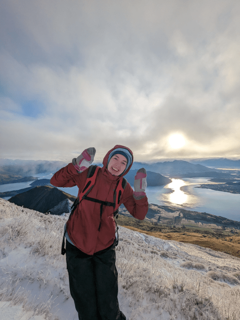 a young white woman wearing winter gear smiles on a mountain top overlooking a body of water