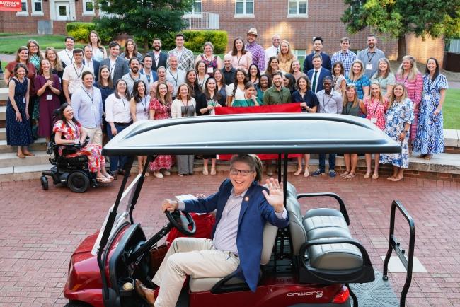 Group of alums and students at the union amphitheater behind president Doug Hicks on a golf cart, waving