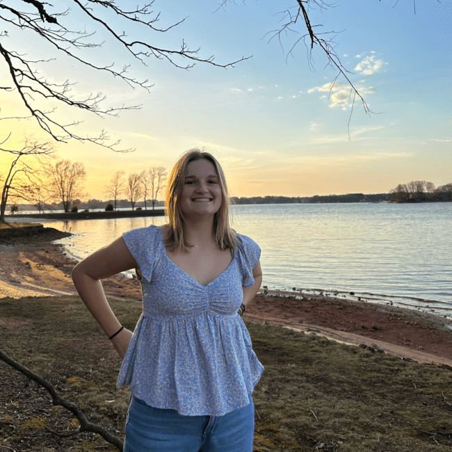 a young white woman standing in front of a lake at sunset