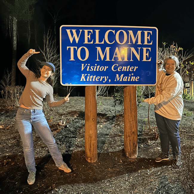 two young white women standing in front of a "Welcome to Maine" sign