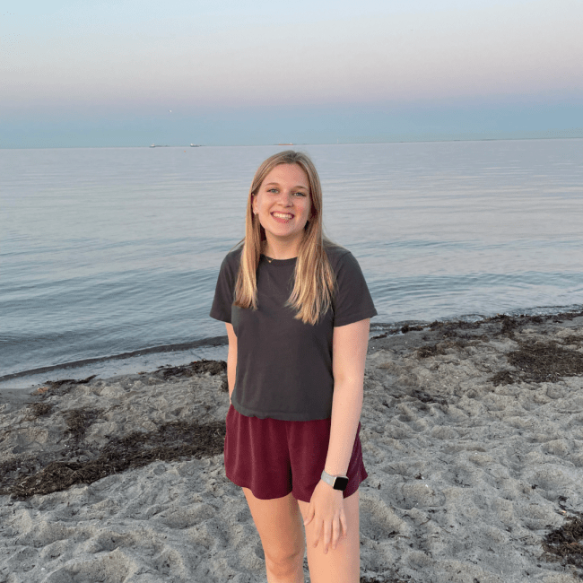 a young white woman standing in front of the ocean at sunset