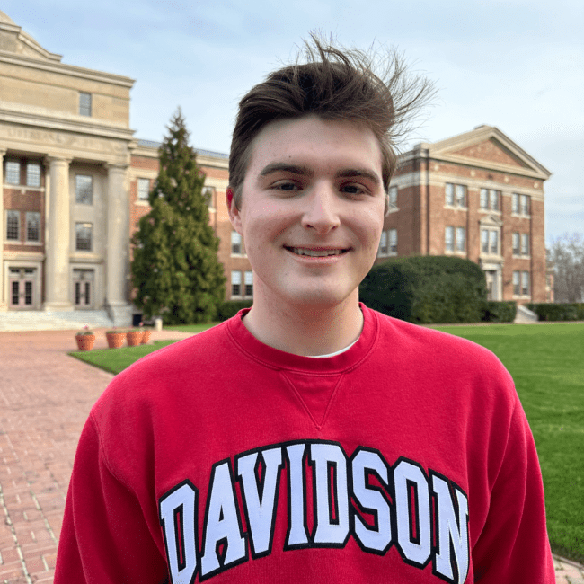 a young white man wearing a Davidson sweatshirt and smiling on campus