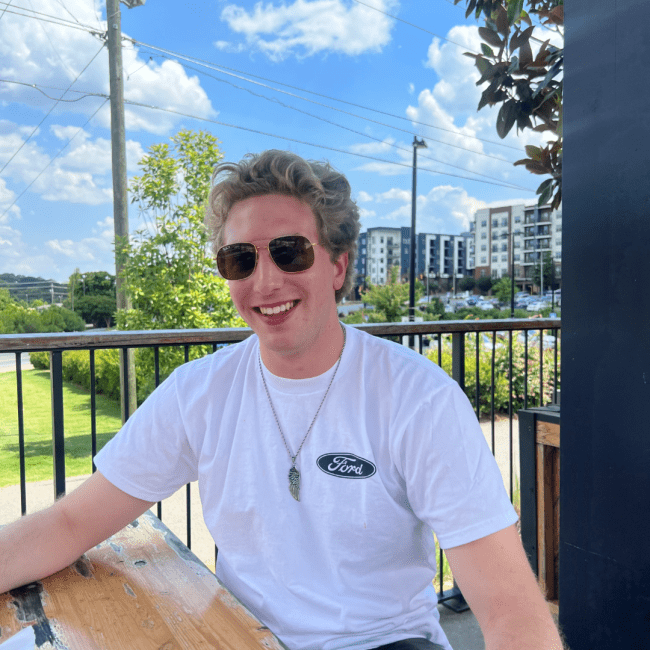 a young white man smiling while sitting at a table outside in a city