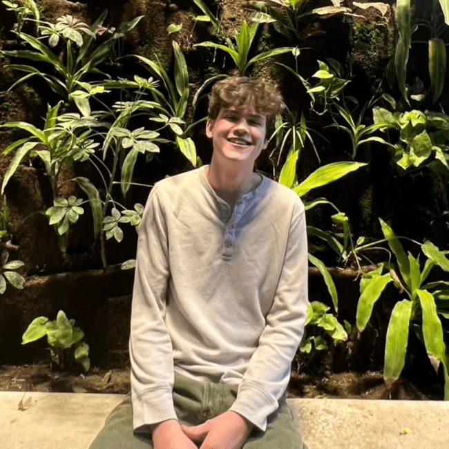 a young white man smiling against a plant wall in background