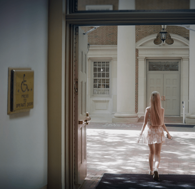 a young white woman with long blonde hair walks outside a building where an accessibility button is clearly visible