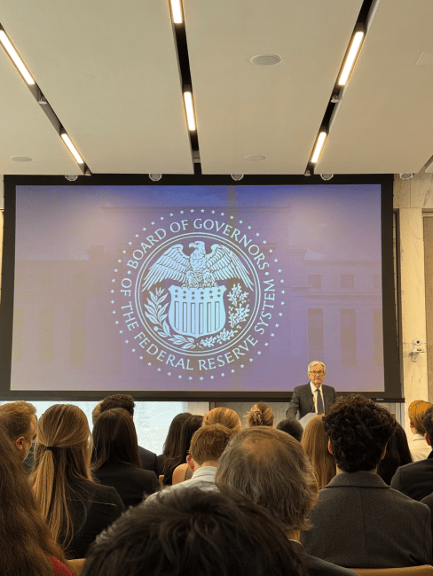 an older white man stands in front of a presentation in a large lecture hall