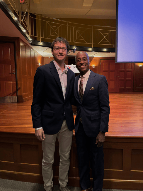 a young white man stands with an older Black man, both wearing suits and ties in an auditorium