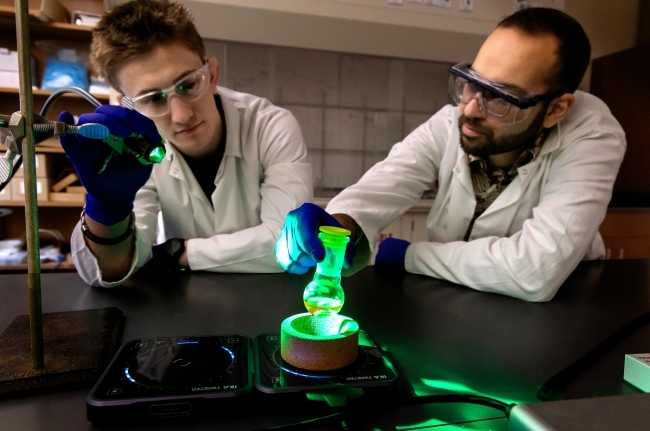 a young white man in a chemistry lab with an older man, both in white lab coats and goggles