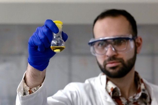 a young man wearing a lab coat and goggles holds up a vial