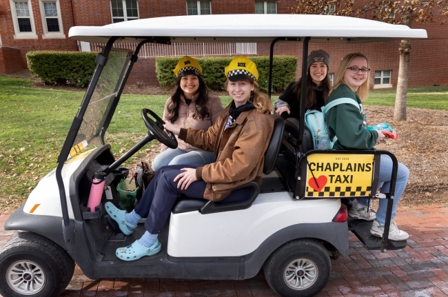 a group of four students sit in a golf cart with a sign that reads "Chaplains' Tax"