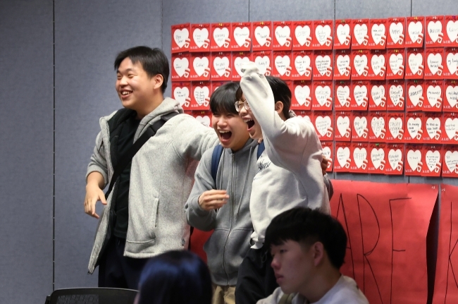 a group of three students dance and smile in front of a wall of heart cards