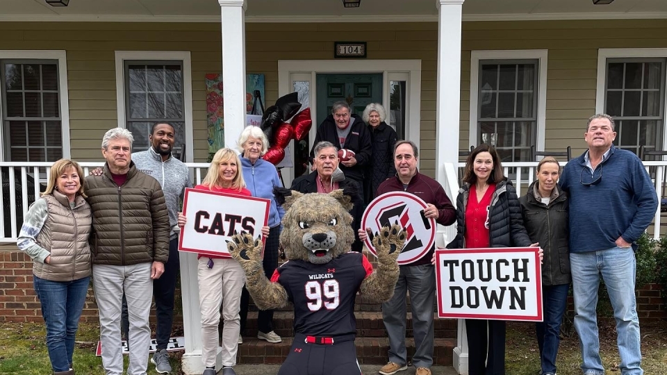 Peter Andrews, group of Davidson staff including president & athletic director, and Lux the wildcat outside Dave & Barbara Fagg's house with Davidson signs  "Cats" and "Touch Down" in hands