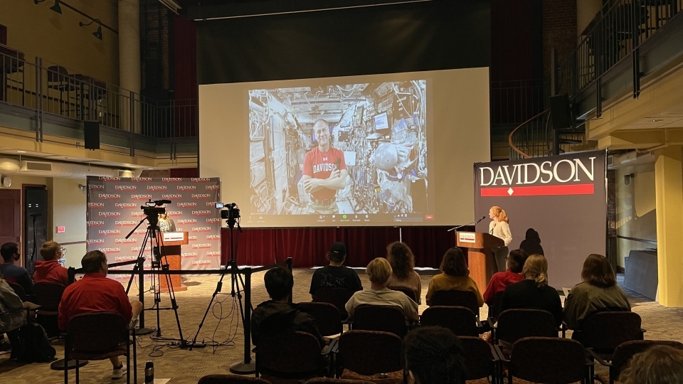 students seated in the 900 room, looking toward the front at Tom Marshburn on a screen in a Davidson t-shirt in a spaceship with two in-person people behind podiums on both sides of the screen