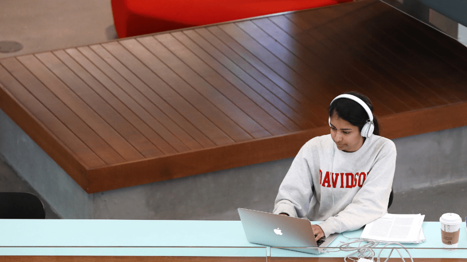 student wearing a Davidson sweatshirt and headphones works on a laptop in a modern academic building