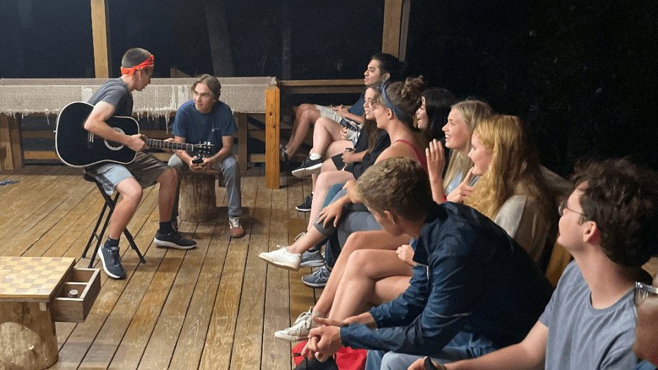 a group of students sit around a cabin as one plays the guitar