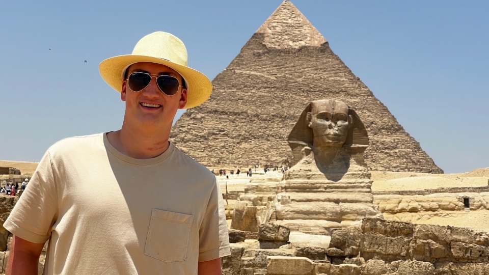 a young white male wears a straw hat while standing in front of an Egyptian pyramid and sphinx