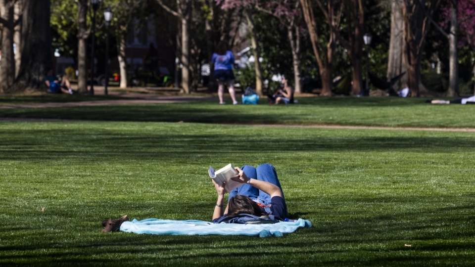 a young woman reads a book while laying on a blanket on a grassy lawn