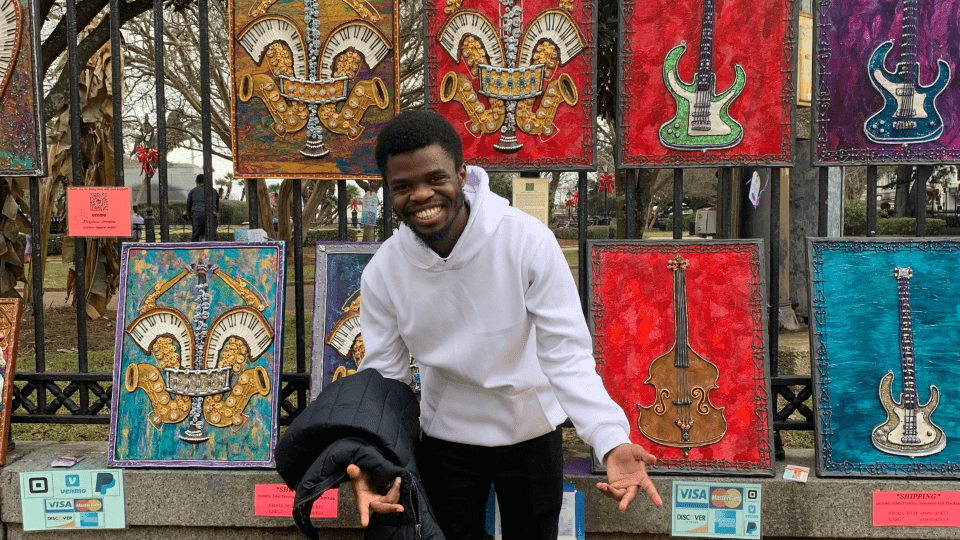 a young Black man wearing a white sweatshirt stands in front of a wall of paintings of musical instruments