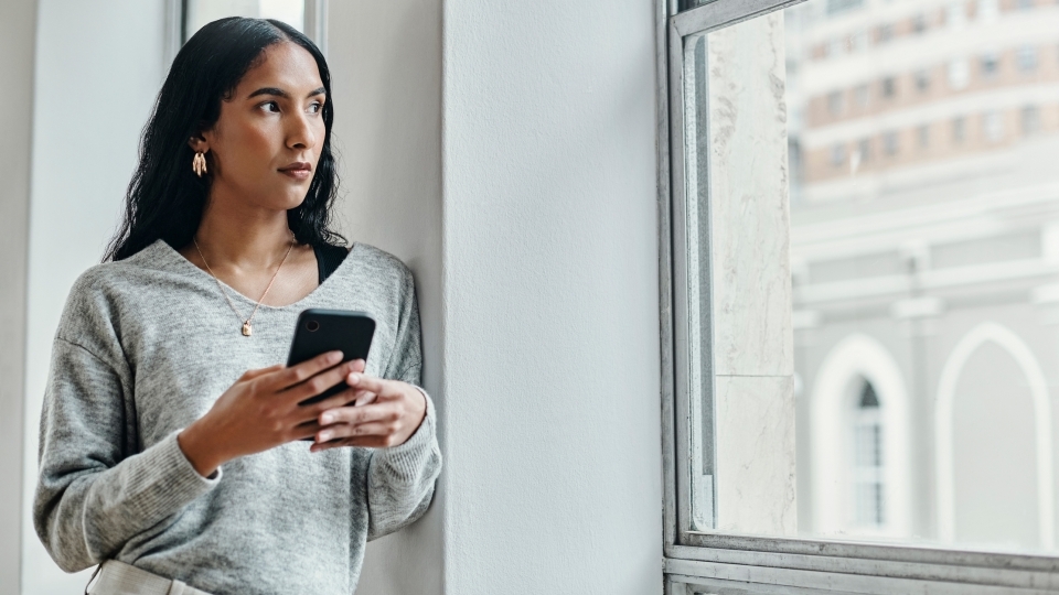 Woman holding a cell phone looking out the window wondering about social media misinformation