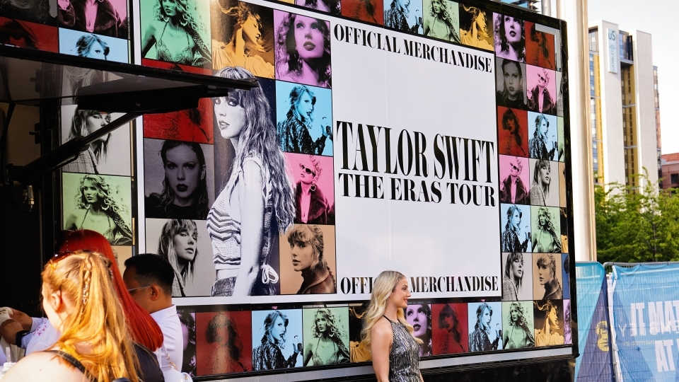 a young woman stands in front of sign that reads "Taylor Swift Eras Tour"