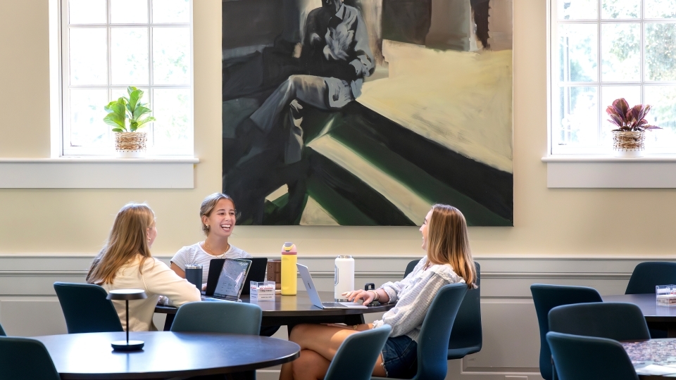three young women smile while gathered around a table with laptops and waterbottles