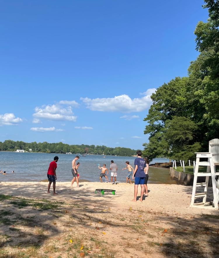 a group of students stand around a beach and swim in a lake on a sunny day