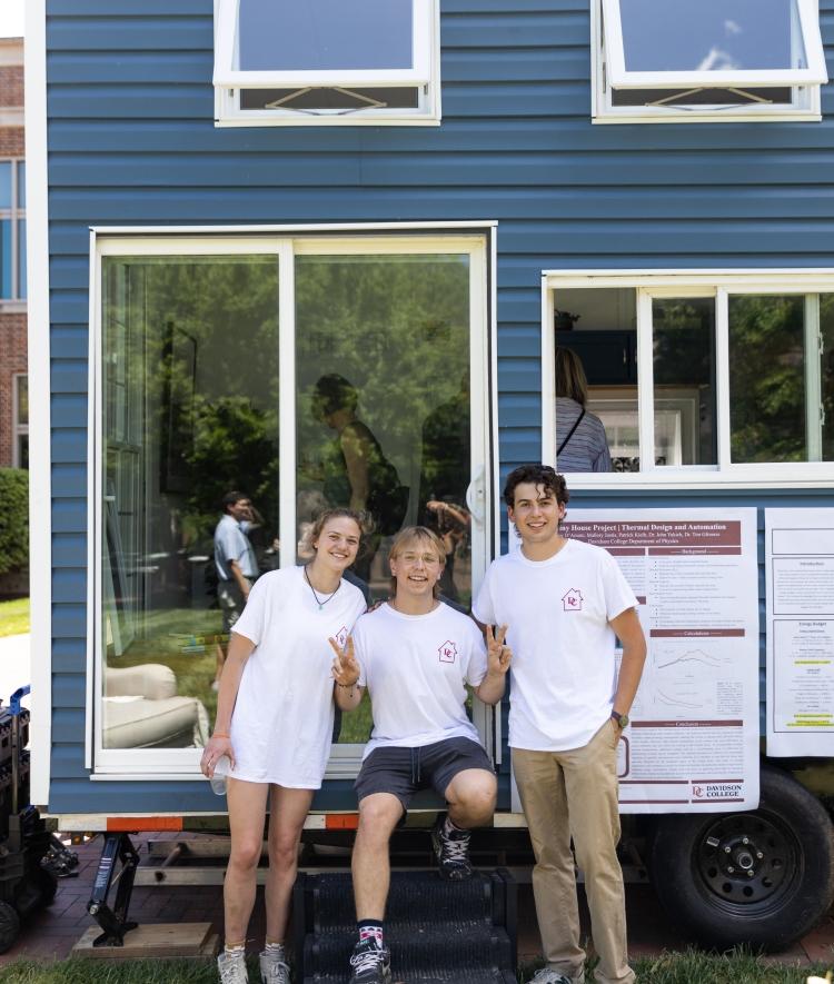 Three students sitting in front of completed tiny house, painted blue with white trim