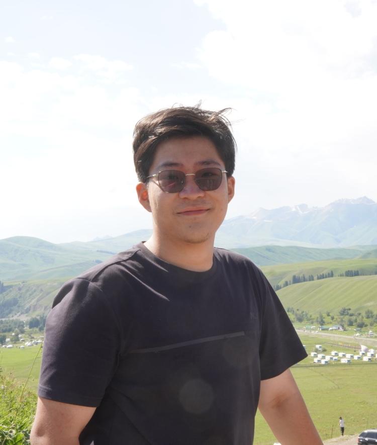 a young Chinese man standing in front of sweeping mountains on a sunny day