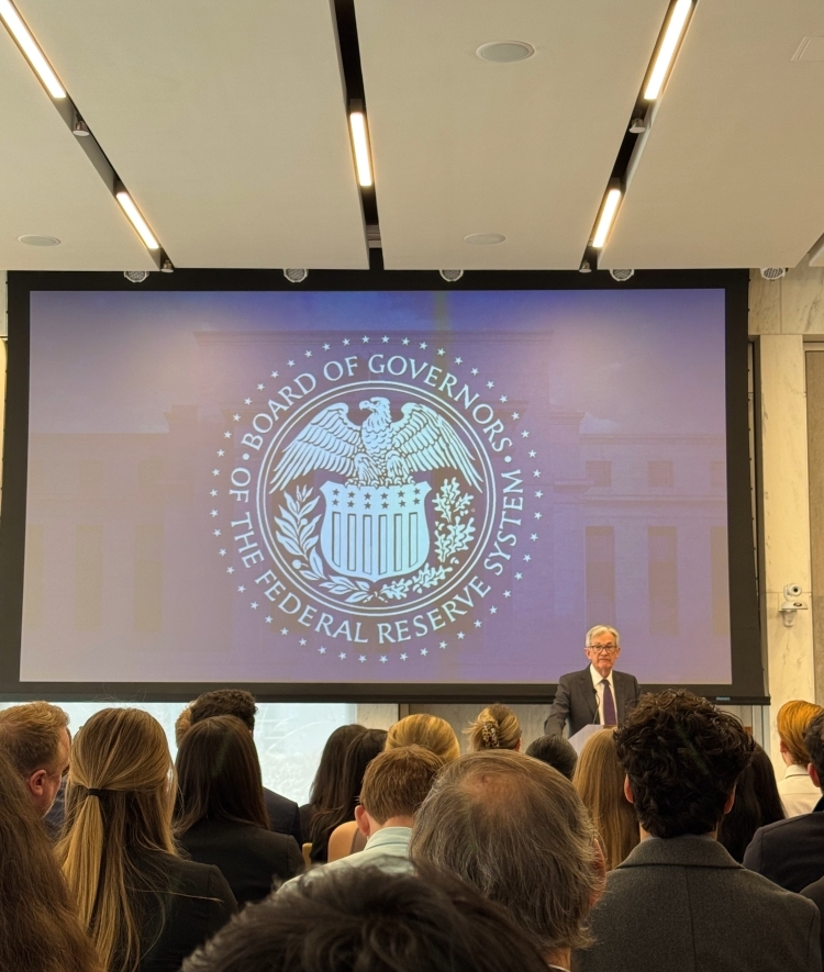 an older white man stands in front of a presentation in a large lecture hall