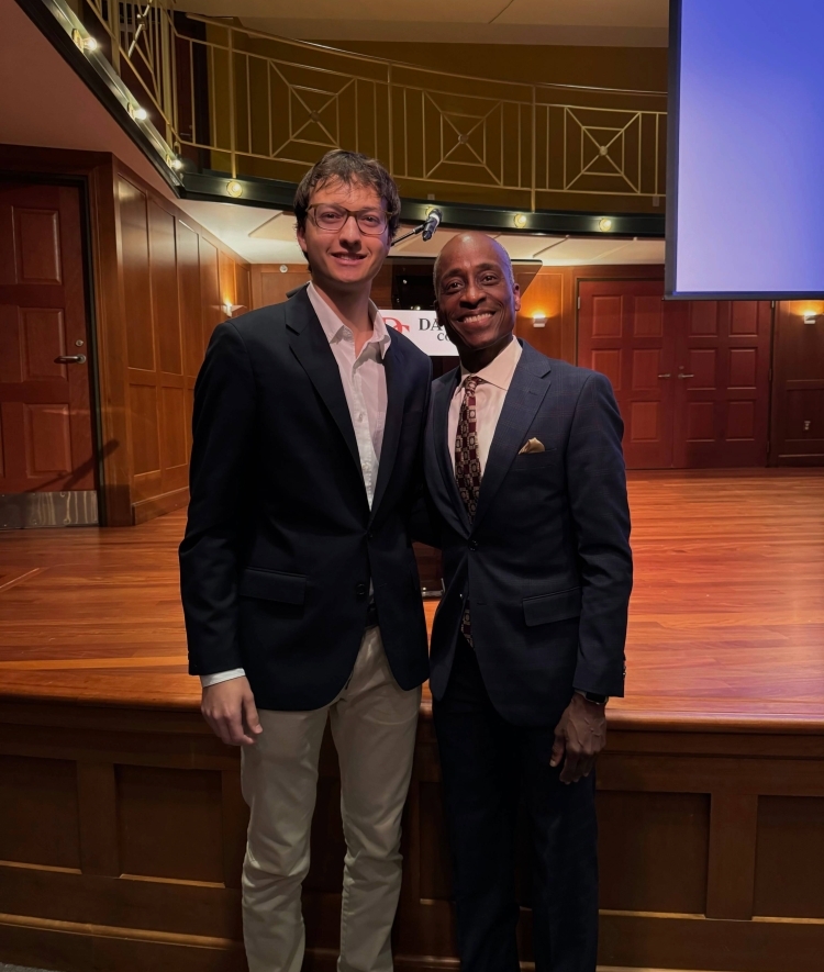 a young white man stands with an older Black man, both wearing suits and ties in an auditorium