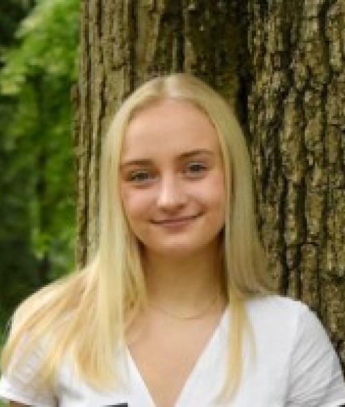 a young white woman with blonde hair wearing a white top standing in front of a tree