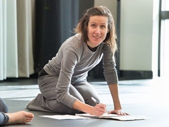 Dance professor sits on studio floor while taking notes and talking to students who are partially visible at the corner of the photo