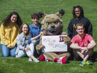 Lux and students sit on grass with sign reading #AllInForDavidson