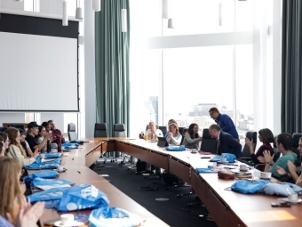 a group of scholars and adults sit around an ovular conference table in a large conference room