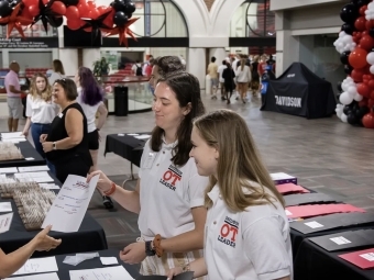 two young women in white polos talk to an older woman around a table with balloons in the background