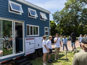 Student team presenting work in front of tiny house, holding microphone