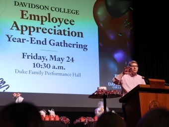 an older man speaks into a podium in front of a screen that reads "Employee Appreciation Year-End Gathering"