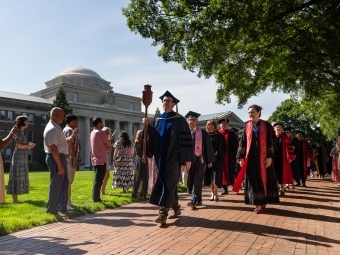 a procession of graduates on a sunny day in front of an academic building