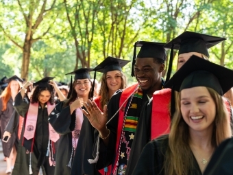 a procession of graduates in graduation regalia smiling on a sunny day outside