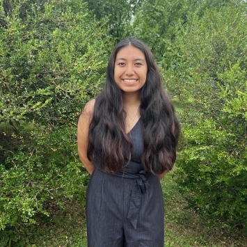 a young woman with brown hair standing in front of greenery