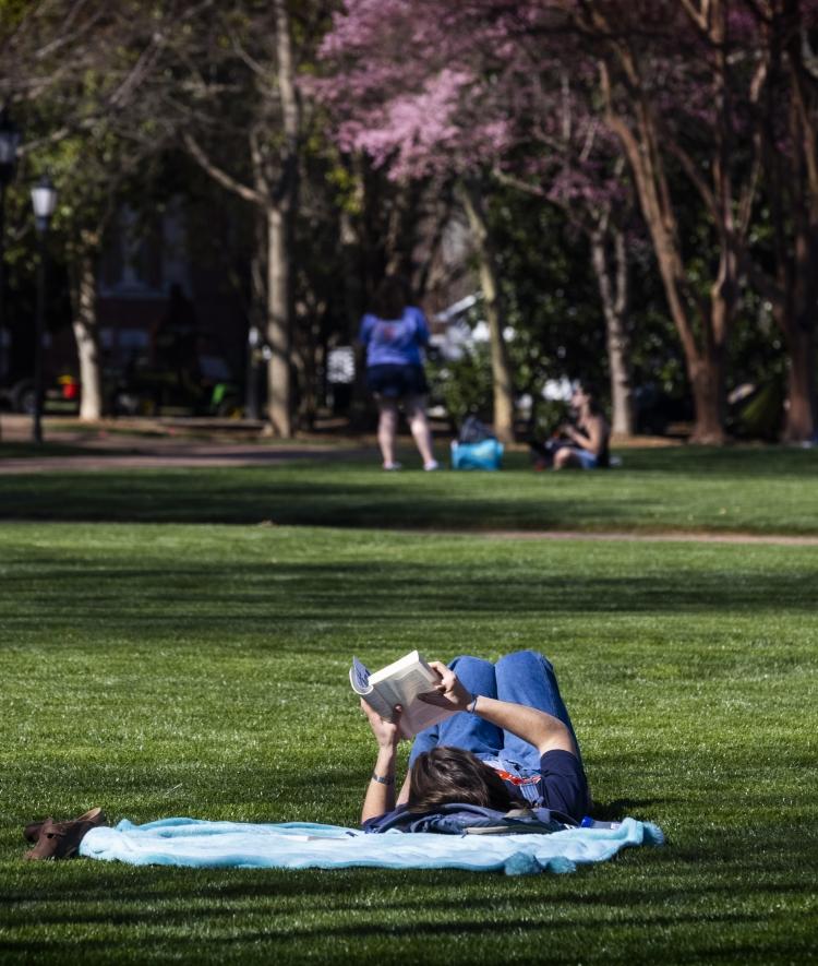 a young woman reads a book while laying on a blanket on a grassy lawn